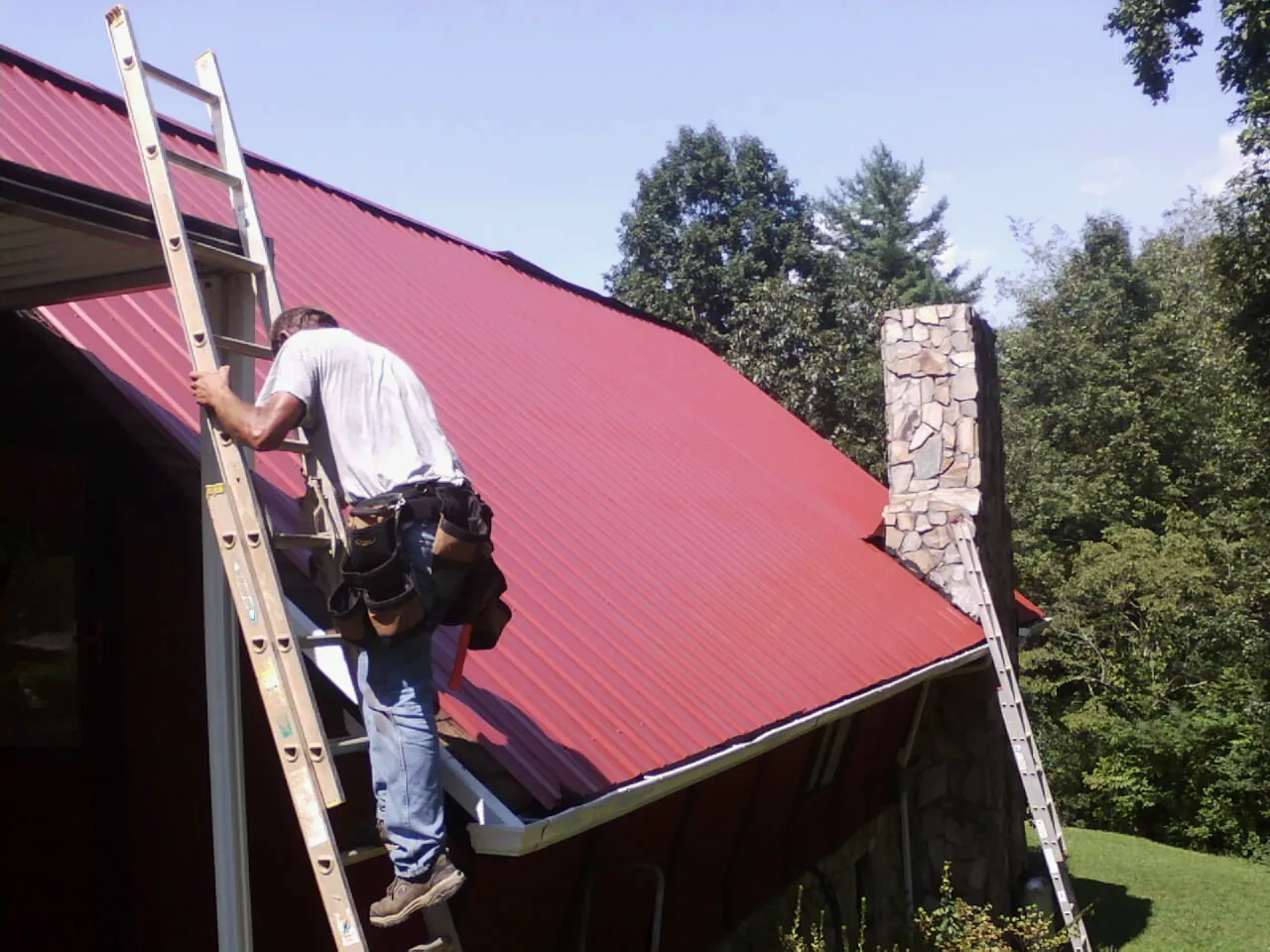 Roofer on a ladder beside a red metal roof and stone chimney — Soffit & Fascia Repair by Roofing Brothers in Merrillville